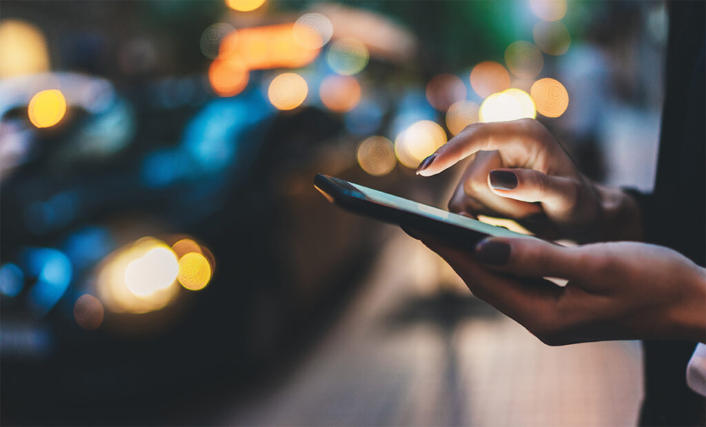 Businesswoman with smartphone using application for calling taxi online walking on city street night, female hands with internet connection texting friend via mobile phone on background bokeh color light